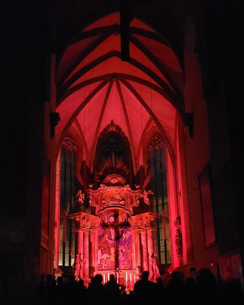 "Der Chorraum von St. Johannis mit der modernen Auferstehungsszene ist rot erleuchtet. Im Vordergrund sieht man Silhouletten von Menschen, die darauf schauen. Das kleine Kreuz auf dem Altar wirft einen riesigen Schatten auf den auferstandenen Christus."