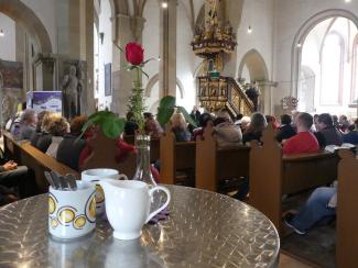 In der Vesperkirche Schweinfurt stehen Milchkännchen und Tische bereit. Die Menschen sitzen noch zum Gottesdienst in den Bänken.